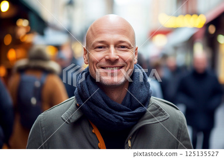 bald man in his 40s or 45 years old, smiling warmly while standing in an autumn cityscape. He wears a cozy scarf and a backpack, dressed for the fall season 125135527