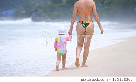 Mother and child stroll along coastline where waves gently meet sand Mother and child stroll along coastline where waves gently meet sand 125138363