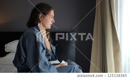 Young caucasian woman in bathrobe sitting on bed holding throat with painful expression Young caucasian woman in bathrobe sitting on bed holding throat with painful expression 125138365