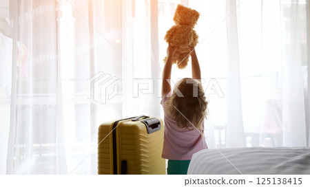 Preschooler girl cuddles cute teddy bear standing near big suitcase 125138415