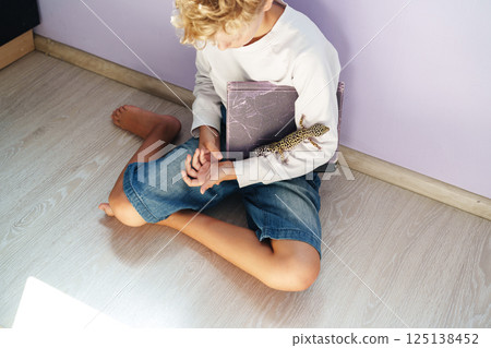 Young boy sits on floor with pet gecko while holding a notebook and enjoying a quiet moment at home Young boy sits on floor with pet gecko while holding a notebook and enjoying a quiet moment at home 125138452