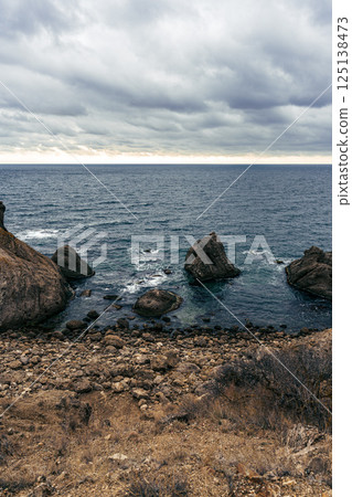 Rocky coastline under a cloudy sky with ocean waves crashing against the shore at dusk 125138473