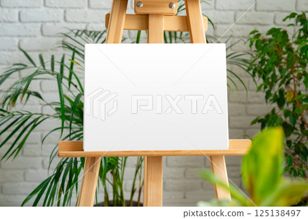 Blank canvas on an easel surrounded by greenery in a bright, modern art studio Blank canvas on an easel surrounded by greenery in a bright, modern art studio 125138497