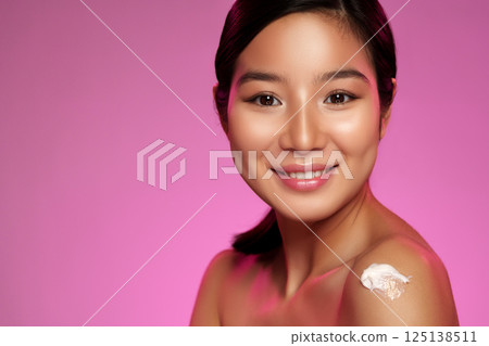 Woman applying skincare cream with a radiant smile in a vibrant pink setting during daylight 125138511