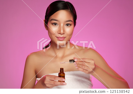 Young woman applies skincare serum in a beauty routine with pink backdrop 125138542