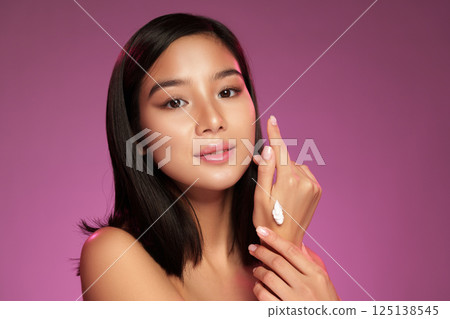 Beautiful woman applying cream on her hand in a styled studio with a purple background during a beauty routine Beautiful woman applying cream on her hand in a styled studio with a purple background during a beauty routine 125138545