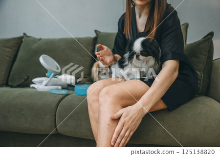 woman applying skincare lotion to her leg while sitting on green couch with two japanese chin dogs during relaxed morning beauty routine, pets calm, skincare, dog lovers, self care brands 125138820