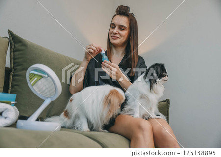 woman applying facial serum with small dropper during morning beauty routine, sitting on green sofa with two japanese chin dogs on her legs woman applying facial serum with small dropper during morning beauty routine, sitting on green sofa with two japanese chin dogs on her legs 125138895