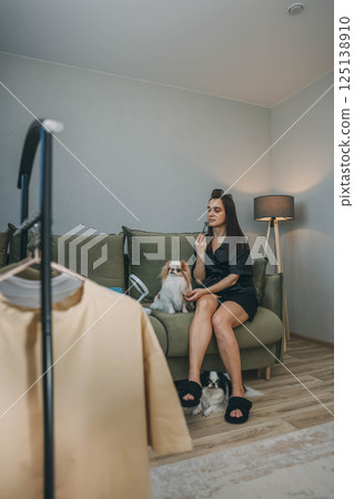 woman in a black bathrobe, relaxing on a green sofa care of her face, massage of face while savoring the company of her two adorable dogs, clothes rack woman in a black bathrobe, relaxing on a green sofa care of her face, massage of face while savoring the company of her two adorable dogs, clothes rack 125138910