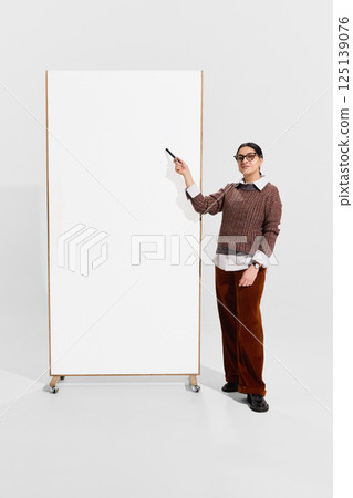 Teacher in brown sweater stands by blank vertical board with marker, confident and composed, prepared for lecture or presentation. 125139076