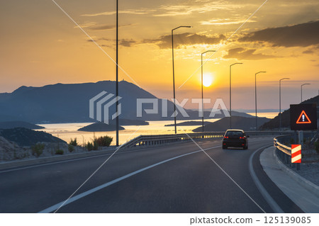 Road on Peljesac peninsula in Croatia, Europe. Scenic sunset drive along coastal highway, mountains in the background. A warning sign cautions for potential hazards. Road on Peljesac peninsula in Croatia, Europe. Scenic sunset drive along coastal highway, mountains in the background. A warning sign cautions for potential hazards. 125139085