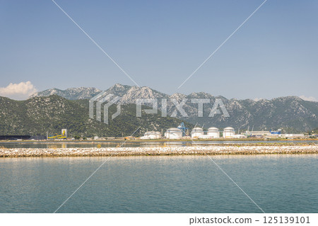 Neretva river near the mouth of river into Adriatic Sea, Croatia, Europe. Coastal Industrial Site: White storage tanks stand against mountains, under a blue sky. Calm water reflects the sunny scene. 125139101