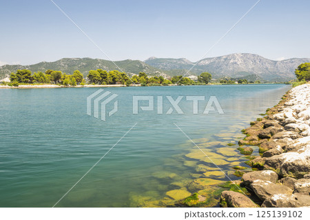 Neretva river near the mouth of river into Adriatic Sea, Croatia, Europe. Scenic waterscape featuring a tranquil river with a rocky shoreline, lush greenery, and distant mountains under a clear sky. 125139102