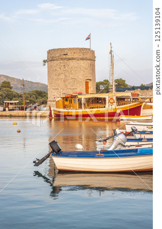 Mali Ston, village in Peljesac peninsula in Croatia, Europe. Historic stone tower overlooks harbor, boats dot the water, Croatian flag waves atop the tower, still calm waters and sunny day. 125139104