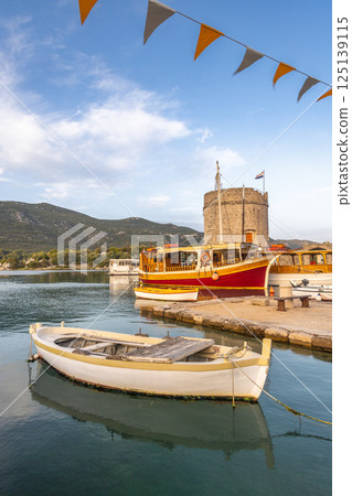 Mali Ston, village in Peljesac peninsula in Croatia, Europe. Picturesque harbor scene with boats, a historic tower, and bunting, showcasing a tranquil maritime atmosphere under a serene sky. Mali Ston, village in Peljesac peninsula in Croatia, Europe. Picturesque harbor scene with boats, a historic tower, and bunting, showcasing a tranquil maritime atmosphere under a serene sky. 125139115