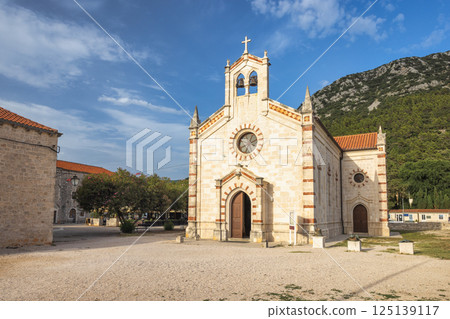 Ston, historic town in Peljesac peninsula in Croatia, Europe. Picturesque church with stone facade and cross-topped bell tower against blue sky near lush mountain landscape. 125139117