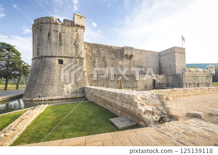 Ston, historic town in Peljesac peninsula in Croatia, Europe. Ancient stone castle with a round tower, surrounded by a moat and grassy area under a bright blue sky with scattered clouds. Ston, historic town in Peljesac peninsula in Croatia, Europe. Ancient stone castle with a round tower, surrounded by a moat and grassy area under a bright blue sky with scattered clouds. 125139118