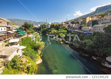 Neretva river in Mostar town, Bosnia and Herzegovina, Europe. Scenic view of a historic town along a river with architecture, lush greenery, and mountains in the backdrop under a clear blue sky. 125139120