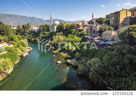 Neretva river in Mostar town, Bosnia and Herzegovina, Europe. Picturesque cityscape featuring a flowing river, historic buildings, and lush greenery, creating a serene and timeless scene. 125139128