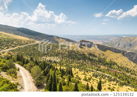 Landscape view from the Fortica hill, Bosnia and Herzegovina, Europe. Scenic view of a winding dirt road through hilly landscape with scattered trees, under a partly cloudy sky. 125139132