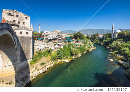 Neretva river near The Old Bridge in Mostar town, Bosnia and Herzegovina, Europe. Scenic view of an old town with a bridge over a river, showcasing historical architecture, serene waters and greenery. 125139137