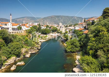 Neretva river in Mostar town, Bosnia and Herzegovina, Europe. Scenic river view with historic architecture, greenery, and a backdrop of mountains. Tranquil cityscape of old town with natural beauty. 125139143