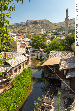 Mostar, historic town in Bosnia and Herzegovina, Europe. Charming riverside town with stone buildings, a bridge, lush greenery, and a minaret against a backdrop of hills and a clear blue sky. 125139144