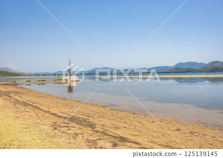Delta of the Neretva river near the mouth of river into Adriatic Sea, Croatia, Europe. Scenic landscape featuring a calm coastline, a small building in water, and distant mountains beneath a blue sky. Delta of the Neretva river near the mouth of river into Adriatic Sea, Croatia, Europe. Scenic landscape featuring a calm coastline, a small building in water, and distant mountains beneath a blue sky. 125139145