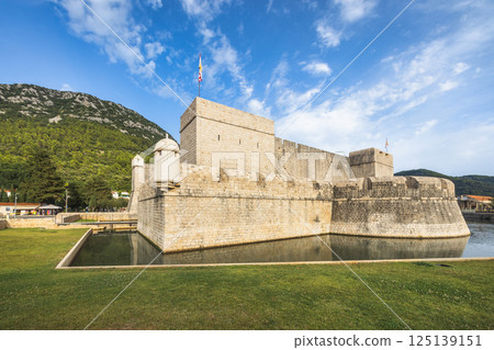 Ston, historic town in Peljesac peninsula in Croatia, Europe. Fortified coastal stone structure reflects in surrounding moat, set against backdrop of lush mountain under sunny blue sky. 125139151