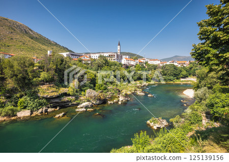 Neretva river in Mostar town, Bosnia and Herzegovina, Europe. Picturesque river landscape featuring a quaint European village nestled among lush greenery under a clear blue sky, conveying tranquility. Neretva river in Mostar town, Bosnia and Herzegovina, Europe. Picturesque river landscape featuring a quaint European village nestled among lush greenery under a clear blue sky, conveying tranquility. 125139156