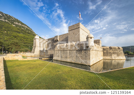 Ston, historic town in Peljesac peninsula in Croatia, Europe. Stone fortress with a moat and flag under a blue sky, against a green mountain backdrop. Tranquil waters reflect the fort. 125139164