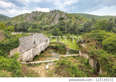 Old stone house in Blagaj village in Bosnia and Herzegovina, Europe. Historic stone building with arched openings over a small river, surrounded by lush greenery and a scenic landscape. 125139172