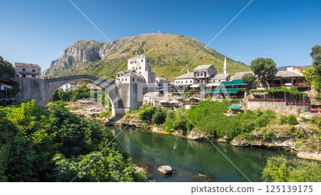 The Stari Most bridge, old bridge on Neretva river in Mostar town, Bosnia and Herzegovina, Europe. Iconic bridge spanning tranquil river with historic buildings, lush greenery, and mountain backdrop. 125139175