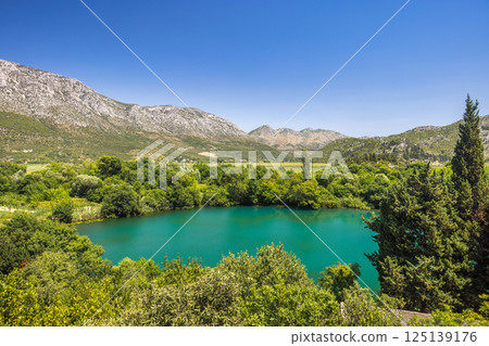 Modro Oko, small lake near delta of the Neretva river, Croatia, Europe. Scenic vista of a turquoise lake reflecting the clear blue sky, framed by lush greenery and majestic mountains in background. 125139176