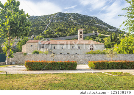 Ston, historic town in Peljesac peninsula in Croatia, Europe. Stone building with a tower near a hillside covered in trees and a path leading to the top, viewed from ground level. Ston, historic town in Peljesac peninsula in Croatia, Europe. Stone building with a tower near a hillside covered in trees and a path leading to the top, viewed from ground level. 125139179