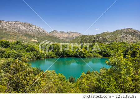 Modro Oko, small lake near delta of the Neretva river, Croatia, Europe. A serene turquoise lake nestled among lush green trees with rolling mountains under a clear blue sky, a peaceful nature scene. Modro Oko, small lake near delta of the Neretva river, Croatia, Europe. A serene turquoise lake nestled among lush green trees with rolling mountains under a clear blue sky, a peaceful nature scene. 125139182