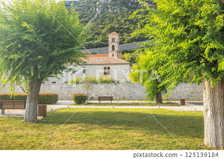 Ston, historic town in Peljesac peninsula in Croatia, Europe. Idyllic view of a park with lush trees and a historic building with a bell tower in the background, creating a serene atmosphere. 125139184