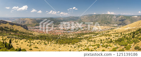 View of Mostar from the Fortica hill, Bosnia and Herzegovina, Europe. Panoramic view of a European city nestled in a valley, surrounded by rugged mountains and clear blue skies. 125139186