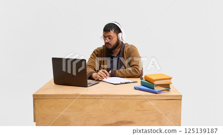 Focused male teacher sitting at desk in headphones, leading online lecture with laptop and colorful books on desk. Focused male teacher sitting at desk in headphones, leading online lecture with laptop and colorful books on desk. 125139187
