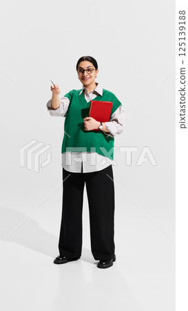Smiling teacher in green vest pointing forward with red notebook in hand during lecture. C Smiling teacher in green vest pointing forward with red notebook in hand during lecture. C 125139188