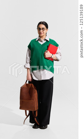 Adult woman as student in green vest standing with backpack and red notebook, ready for university with calm expression. Adult woman as student in green vest standing with backpack and red notebook, ready for university with calm expression. 125139189