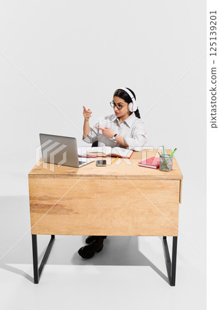 Confident woman in white headphones pointing at laptop screen while teaching online with open books on desk. 125139201