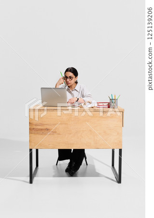 Focused woman, teacher in white blouse sitting at desk with stationary and book, working on laptop and preparing for the lessons on white background. 125139206