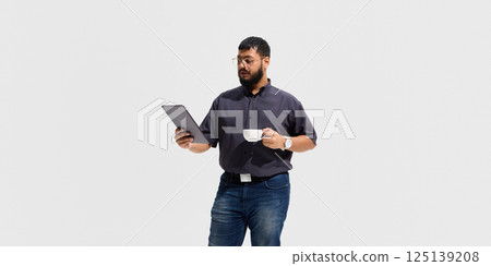 Calm male educator holding coffee cup and tablet, reading attentively, symbolizing thoughtful analysis, digital literacy and relaxed professionalism 125139208