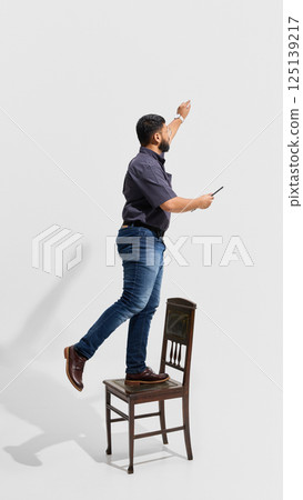 Male teacher in jeans and short-sleeved shirt standing on chair and reaching up, demonstrating active teaching method, engagement and determination 125139217