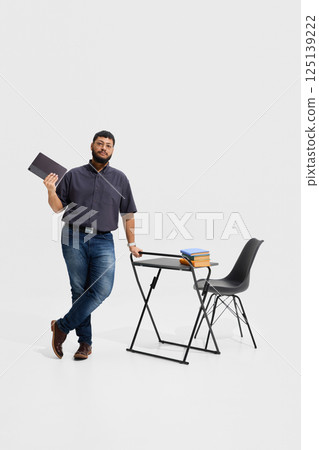 Focused man, teacher leaning on desk with tablet and stack of books, showing relaxed yet professional attitude in classroom Focused man, teacher leaning on desk with tablet and stack of books, showing relaxed yet professional attitude in classroom 125139222