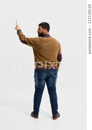Male teacher in brown cardigan raising hand with stylus while facing board, representing engagement and use of technology in classroom 125139226