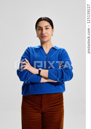 Portrait of focused woman, teacher in blue knitwear crossing arms and standing firmly, conveying leadership, certainty and academic discipline 125139227