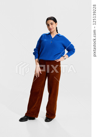 Confident female educator in blue sweater and brown pants standing with hand on hip, radiating authority, readiness and professional engagement 125139228