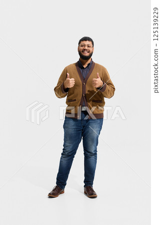 Cheerful male teacher in brown cardigan and jeans giving double thumbs up with confident smile, expressing support 125139229